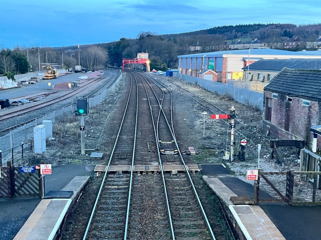 The barrow crossing at Hexham station at twilight. A green signal can be seen with a semaphore signal on the other side of the tracks. The old signal box can be seen in the distance.