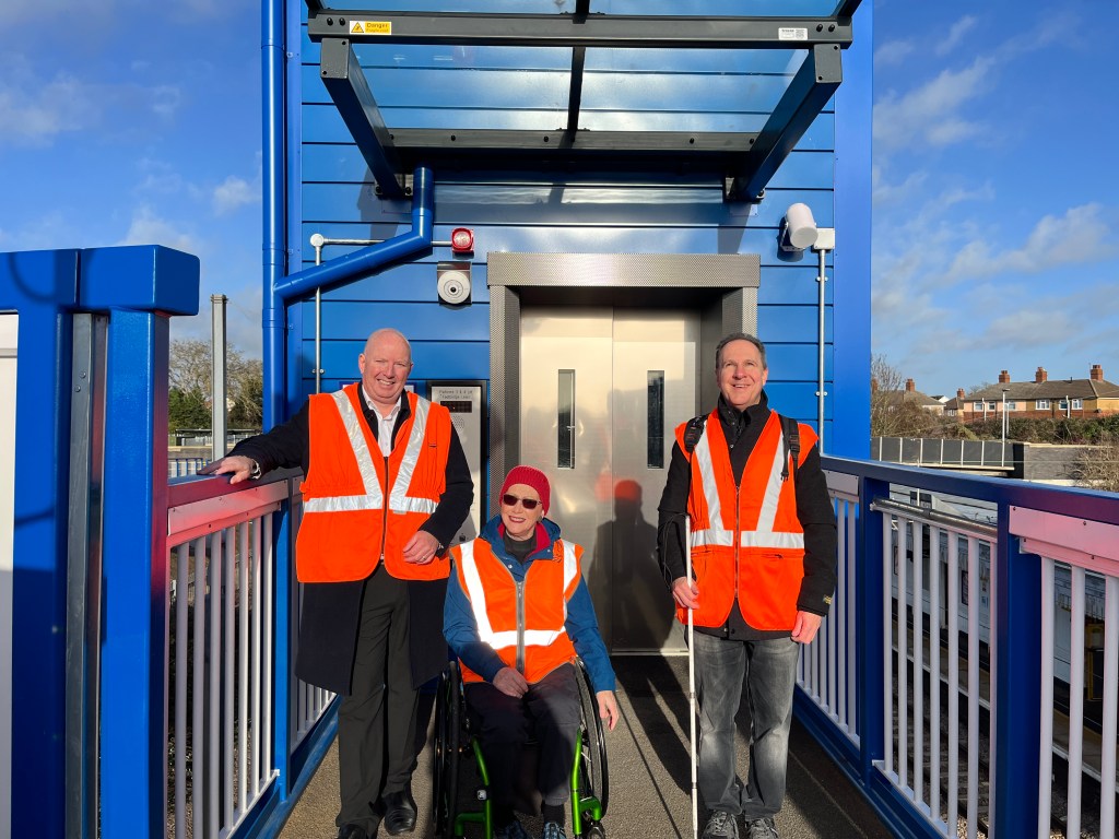 A shot of Julian, Fiona and Paul standing in front of one of the new lifts at Biggleswade station. The lift surround and railings are blue and the background is a blue sky.